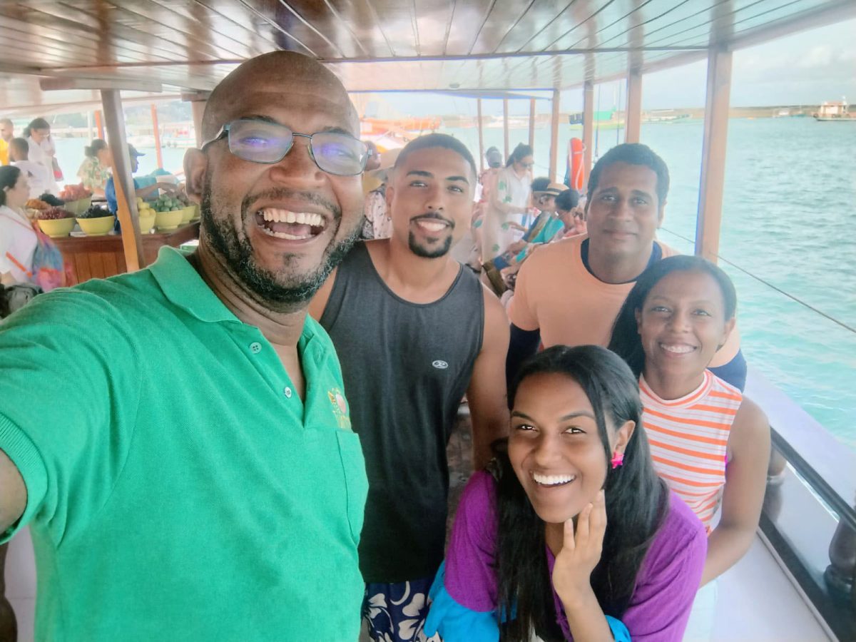 Um grupo de cinco pessoas felizes tirando uma selfie a bordo de uma escuna, com um homem de camisa verde na frente, possivelmente o guia turístico, e outros passageiros ao fundo.