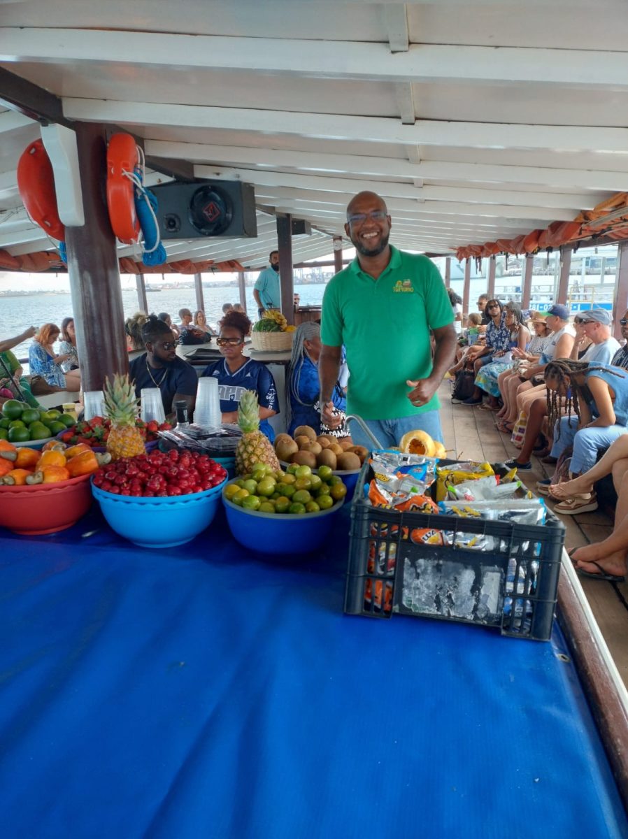 Guia turístico de camisa verde com um sorriso largo em pé ao lado de uma mesa cheia de frutas tropicais e bebidas, com passageiros ao fundo em uma escuna.