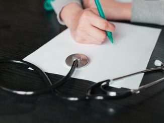 Uma mão segurando um lápis sobre um papel branco ao lado de um estetoscópio sobre uma mesa escura.