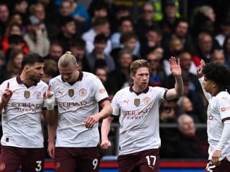 Quatro jogadores do Manchester City comemoram um gol durante uma partida, com expressões de alegria e gestos de vitória.