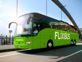 Um ônibus verde da FlixBus parado em uma estação rodoviária, com o sol brilhando ao fundo e a silhueta de uma cidade e veleiros.