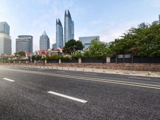 Vista de uma rua vazia com uma faixa de pedestres em primeiro plano, cercada por edifícios modernos de vidro e metal contrastando com construções tradicionais de telhados vermelhos ao fundo, sob um céu claro.