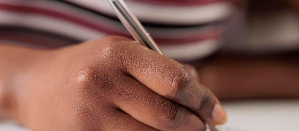 Imagem de uma pessoa concentrada escrevendo em um caderno, com um close-up em sua mão segurando uma caneta azul.