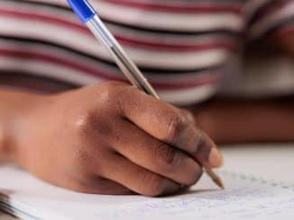 Imagem de uma pessoa concentrada escrevendo em um caderno, com um close-up em sua mão segurando uma caneta azul.