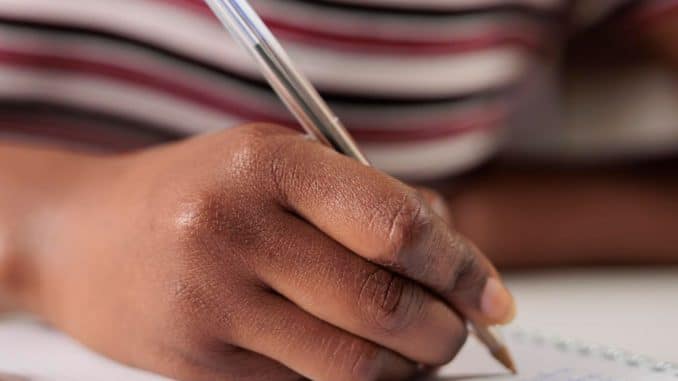 Imagem de uma pessoa concentrada escrevendo em um caderno, com um close-up em sua mão segurando uma caneta azul.