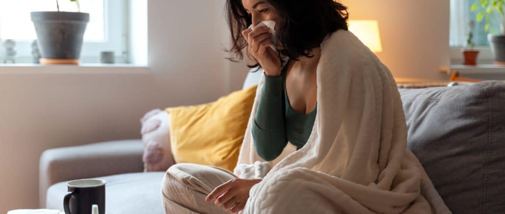 Mulher sentada no sofá enrolada em um cobertor branco, assoando o nariz com um lenço de papel, com uma caneca, óculos e medicamentos ao seu lado, indicando um dia de descanso por motivo de saúde.