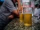Dois copos de cerveja sobre um balcão de bar, com um homem ao fundo, sugerindo o consumo de bebidas alcoólicas e os efeitos desse hábito na sociedade.