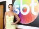 Virginia Fonseca sorrindo, usando vestido amarelo claro, posando à frente do logotipo colorido do SBT.