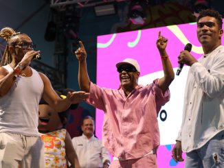 Cantores negros no palco. O artista ao centro veste rosa e levanta os braços, ladeado por outros dois músicos.