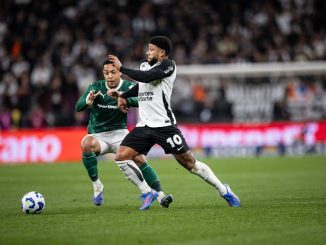 Jogador do Corinthians com camisa branca disputa bola com jogador do Palmeiras de verde em estádio lotado.