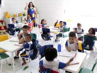 Crianças pequenas sentadas em mesas coloridas realizando atividades escolares em uma sala de aula de educação infantil.