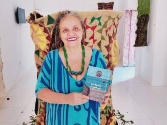 A artista visual Gleciara Ramos sorri em meio às suas obras de arte têxtil na galeria do museu.